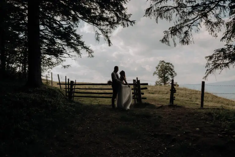 Brautpaar steht silhouettenhaft im Wald, im Hintergrund eine Lichtung und ein Holzzaun